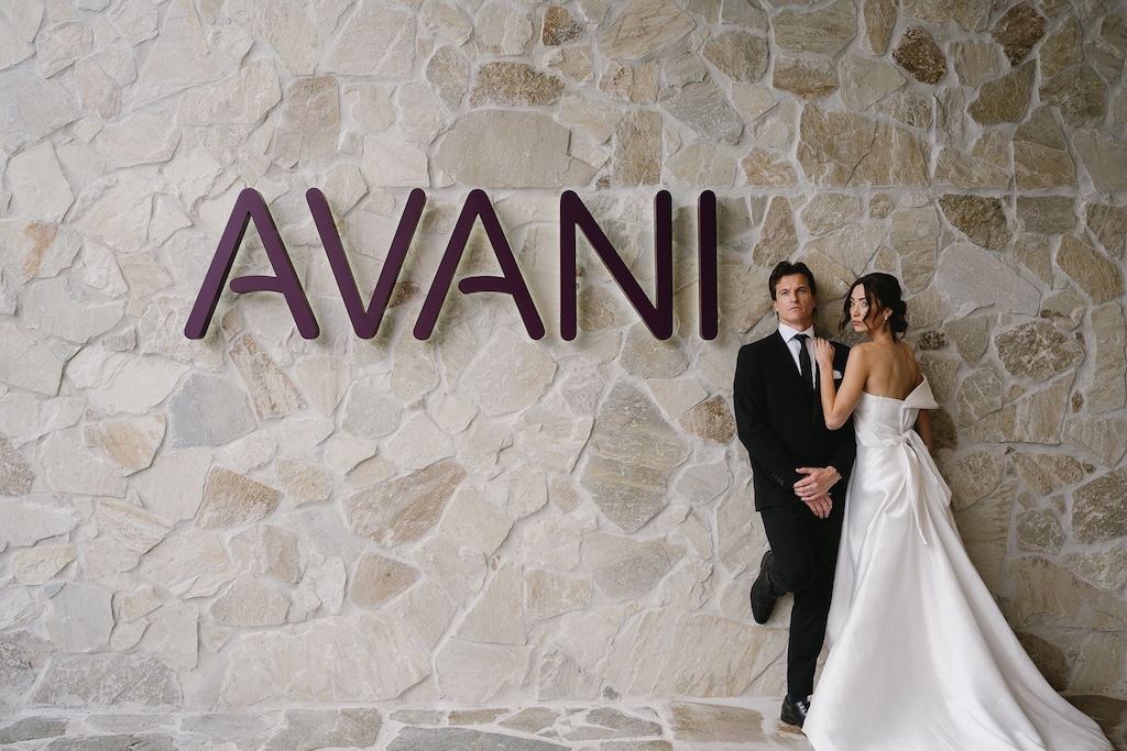 Bride and groom posing beside the illuminated AVANI sign on a stone wall at Avani Mooloolaba Beach Hotel